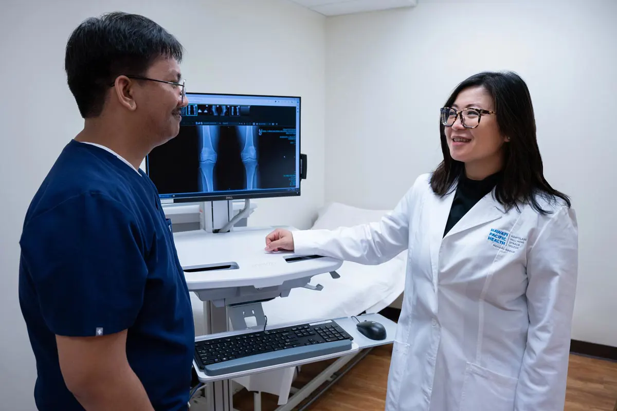 A male medical assistant and female doctor review an X-ray of a knee in an exam room.