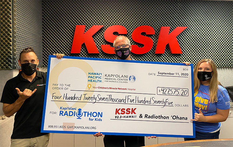Three masked people holding a giant check for $427K in the KSSK radio studio.