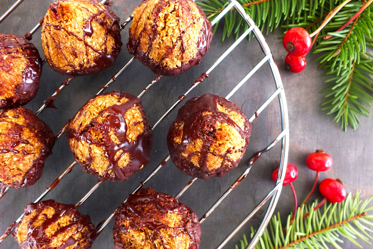 Dark Chocolate-Dipped Mint Macaroon cookies rest on a wire rack on a slate counter top decorated with evergreen branches and holly.