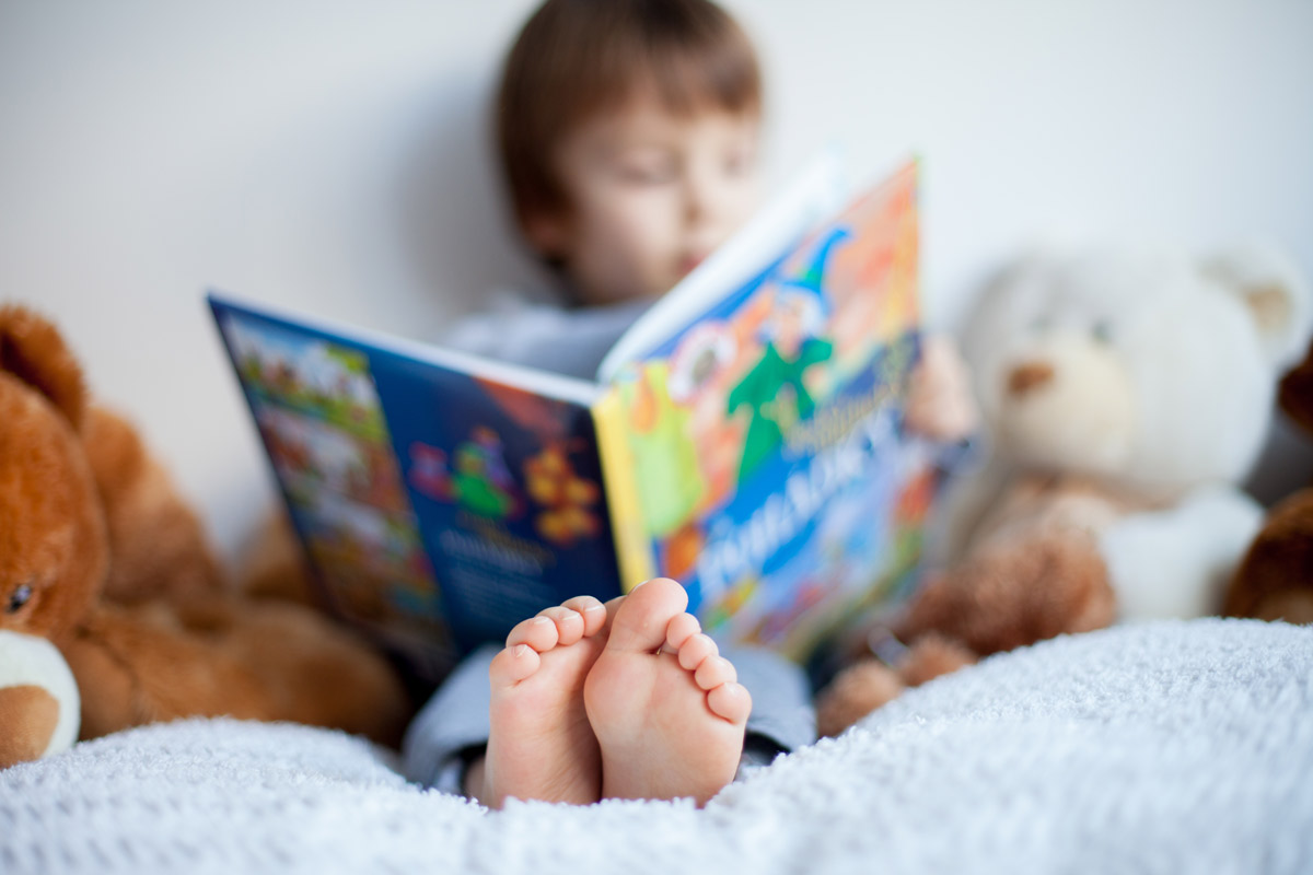 A young boy reads a book in bed surrounded by stuffed animals.