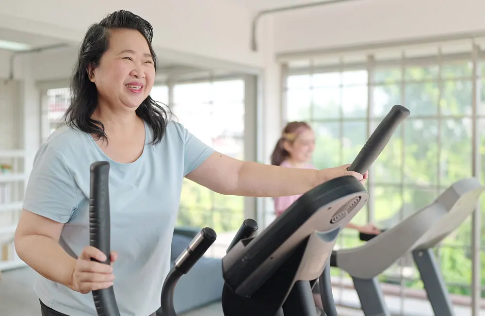 a woman smiles as she exercises on an elliptical machine in a brightly lit exercise room.