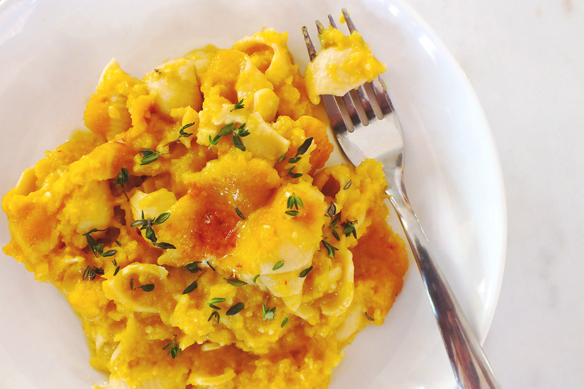 Bright yellow butternut squash mac and cheese with thyme garnish sits on a white plate with a fork.