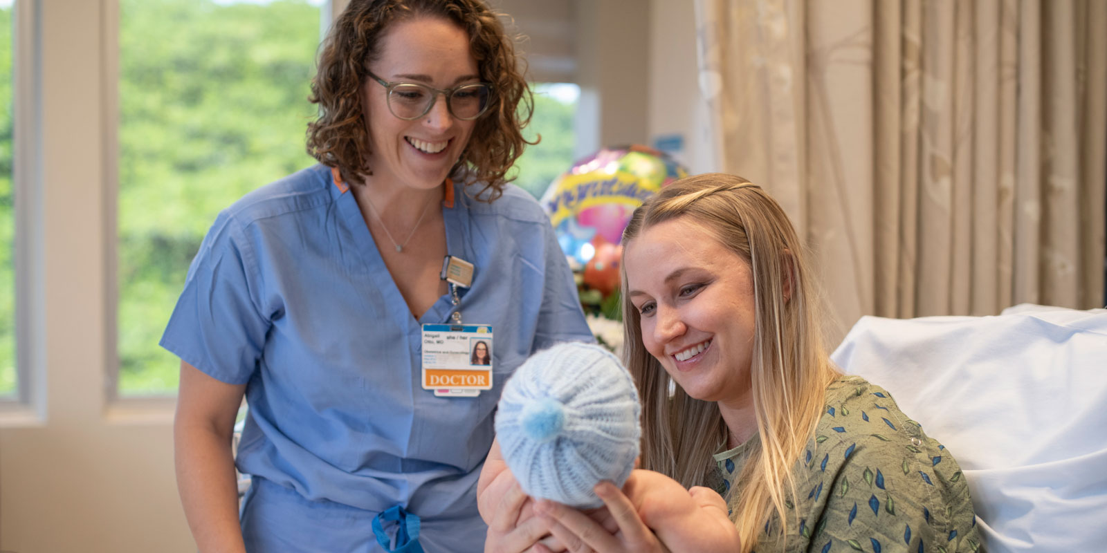 Postpartum mother smiles at the newborn she's holding as a smiling doctor looks on.