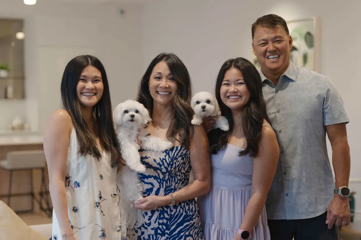 a woman and man smile at the camera as they stand with their two adult daughters and two small, white dogs in their home