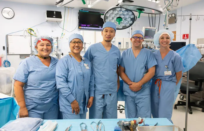 Surgeons in blue scrubs pose and smile at the camera in an operating room.