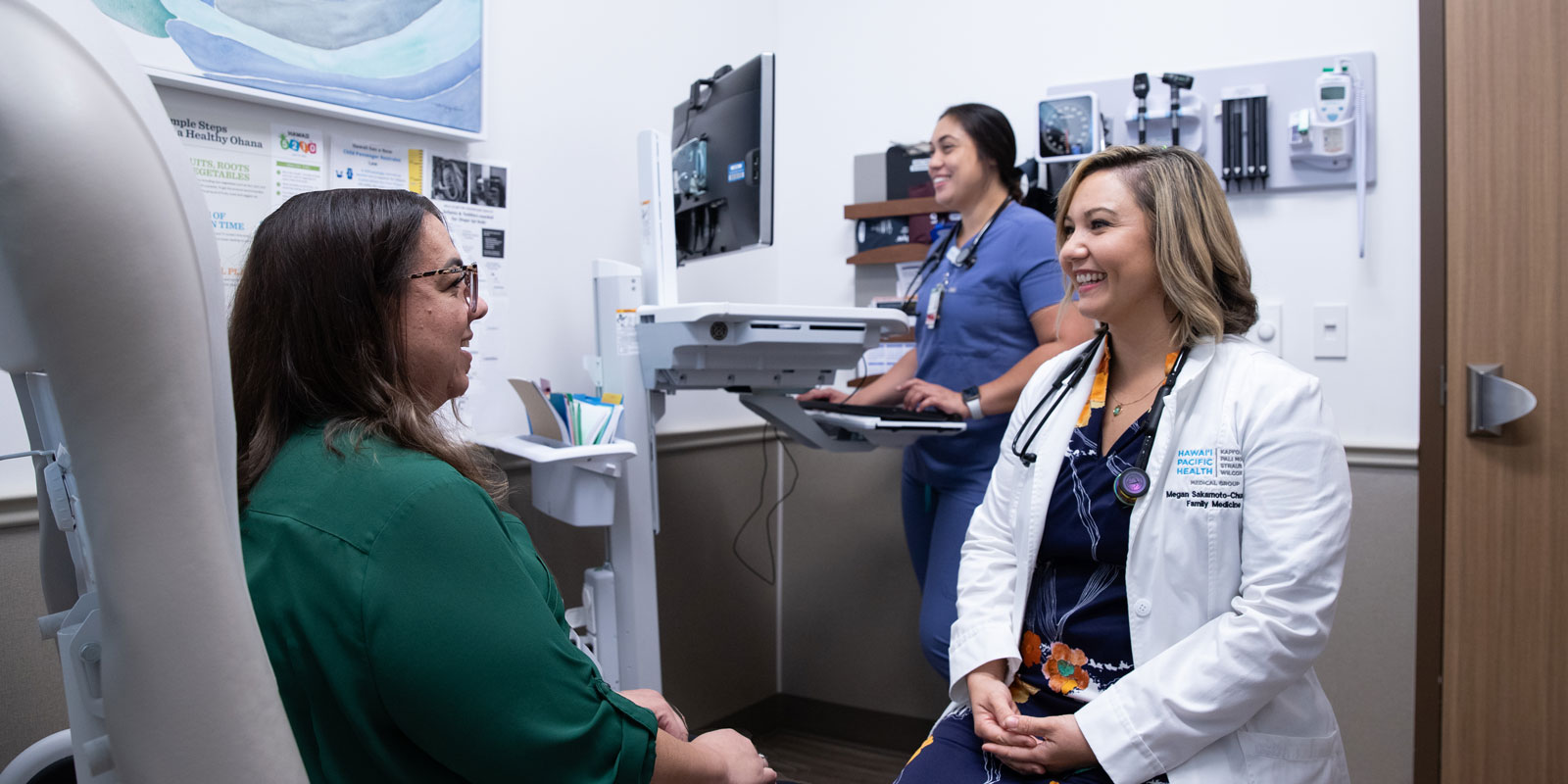 a female doctor smiles while talking with a female patient in an exam room as a female MA types in information to the patient's chart in the background.