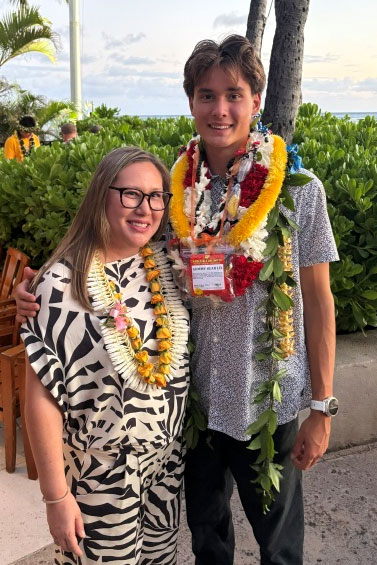 Woman and young, tall man standing together at an outdoor event, both wearing lei. 