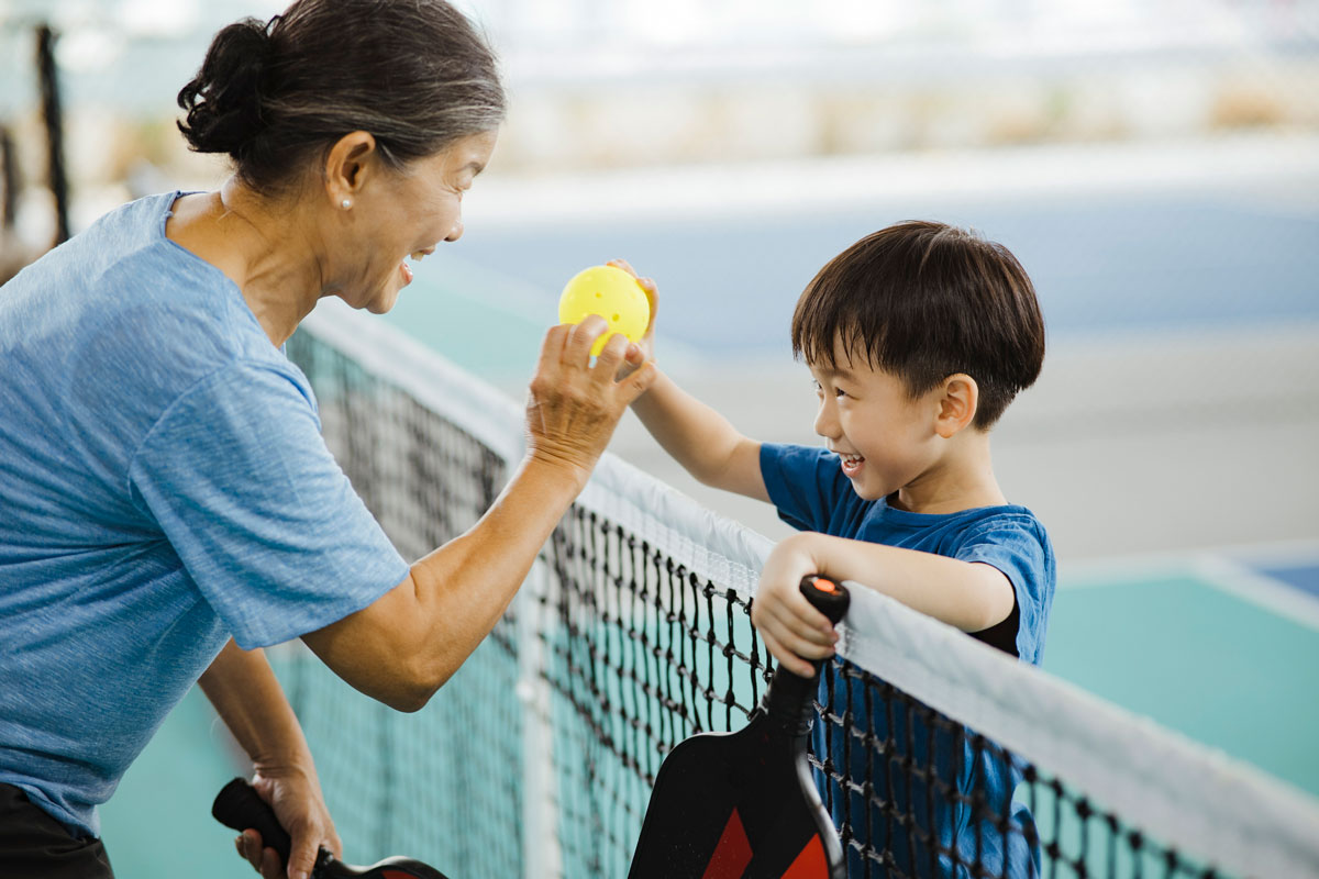 a grandmother hands a pickleball to her grandson over the net on a pickleball court