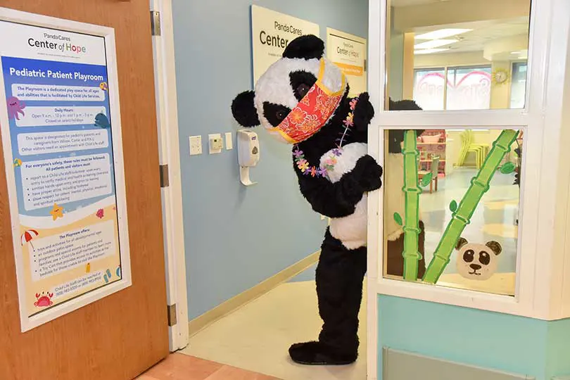Person in a masked panda costume playfully posing by the Panda Cares Center of Hope entrance.