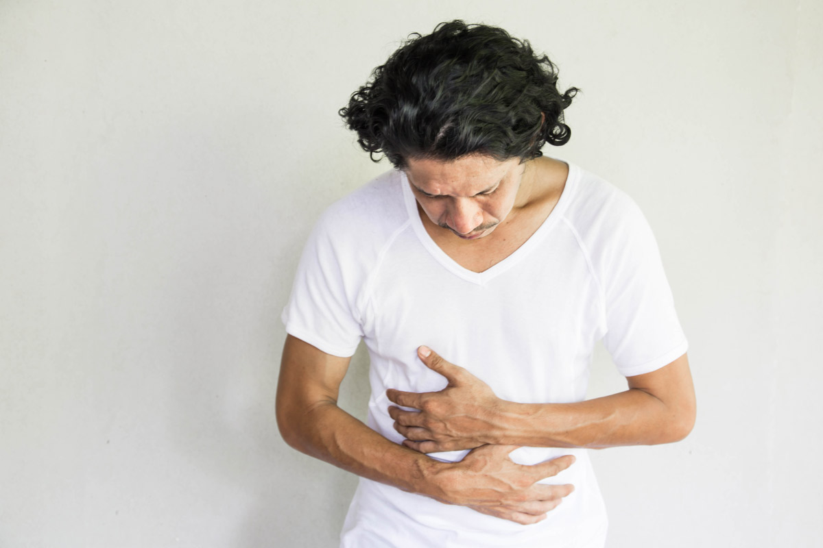 A middle-aged man in a white T-shirt holds his stomach with a look of pain on his face to indicate possible colon cancer.