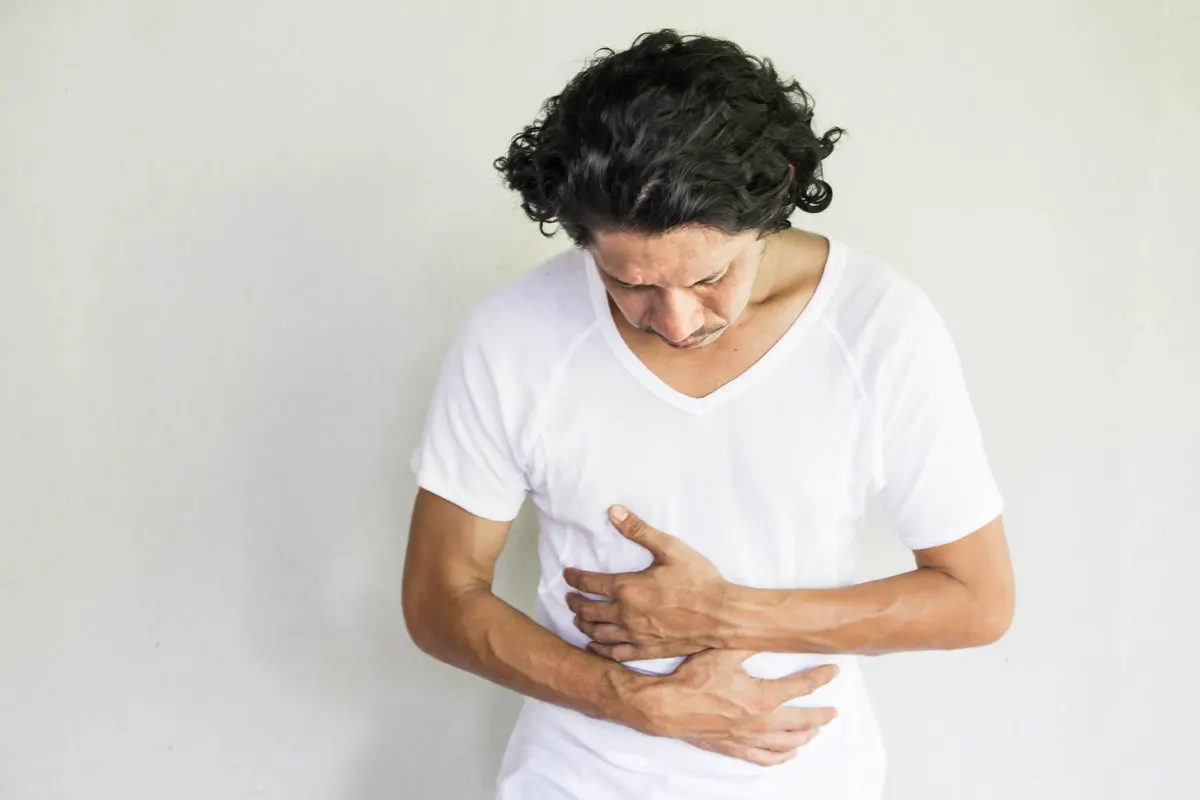 A middle-aged man in a white T-shirt holds his stomach with a look of pain on his face to indicate possible colon cancer.