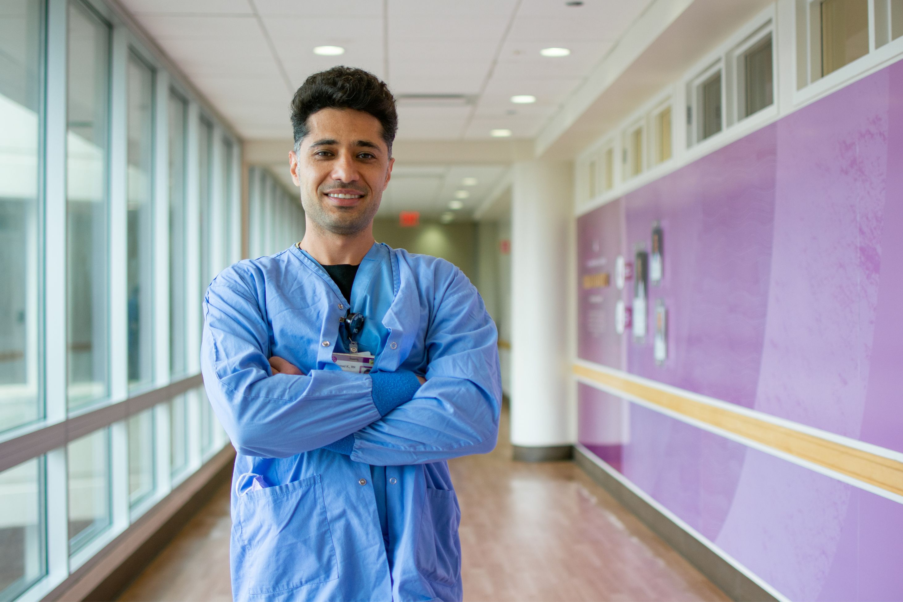 Healthcare worker in scrubs stands in a hospital hallway with arms crossed.