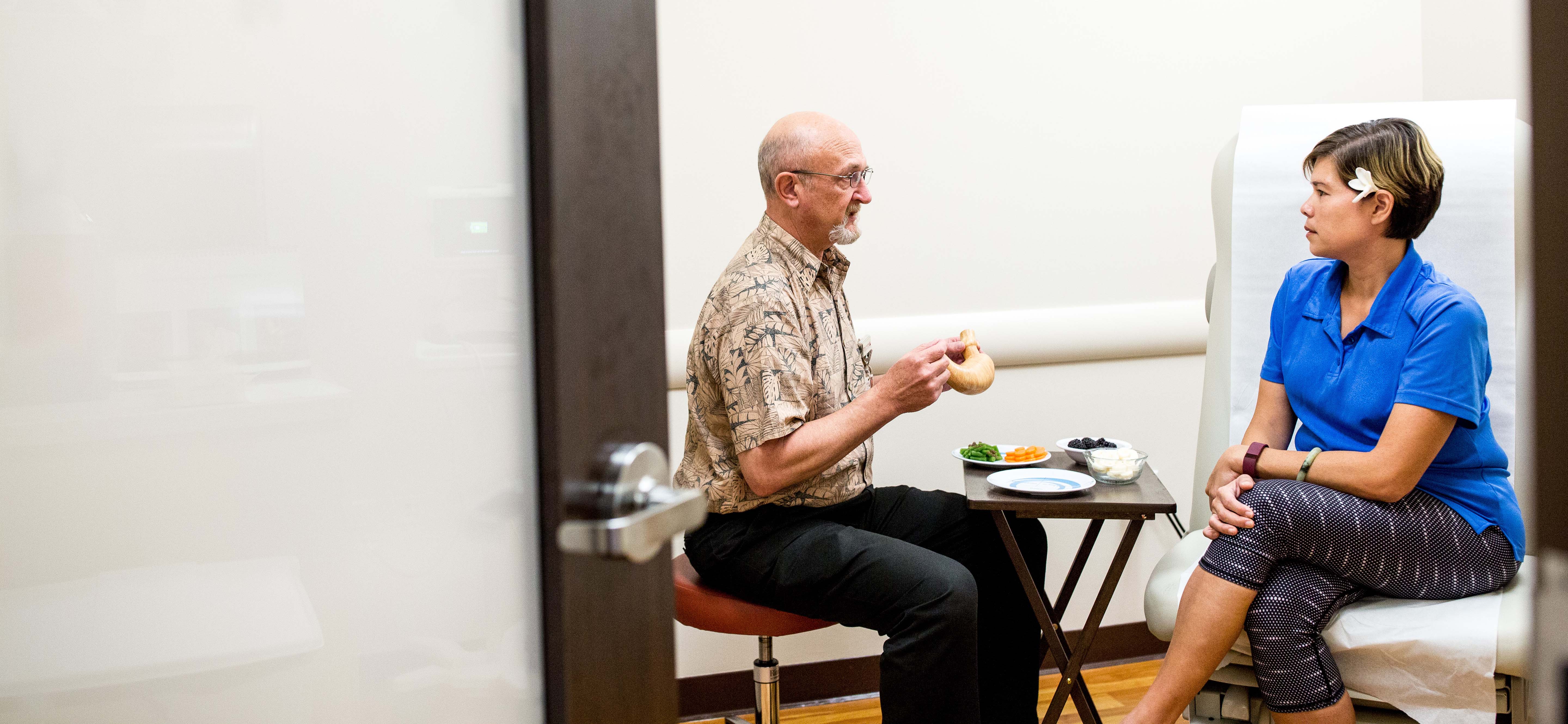 Two people sit talking with some fruits and vegetables on a table nearby.