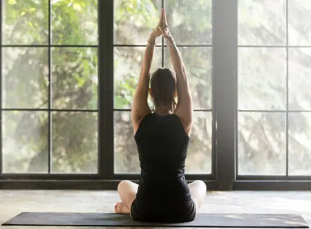 A person practices a seated yoga pose in front of a wall of windows.