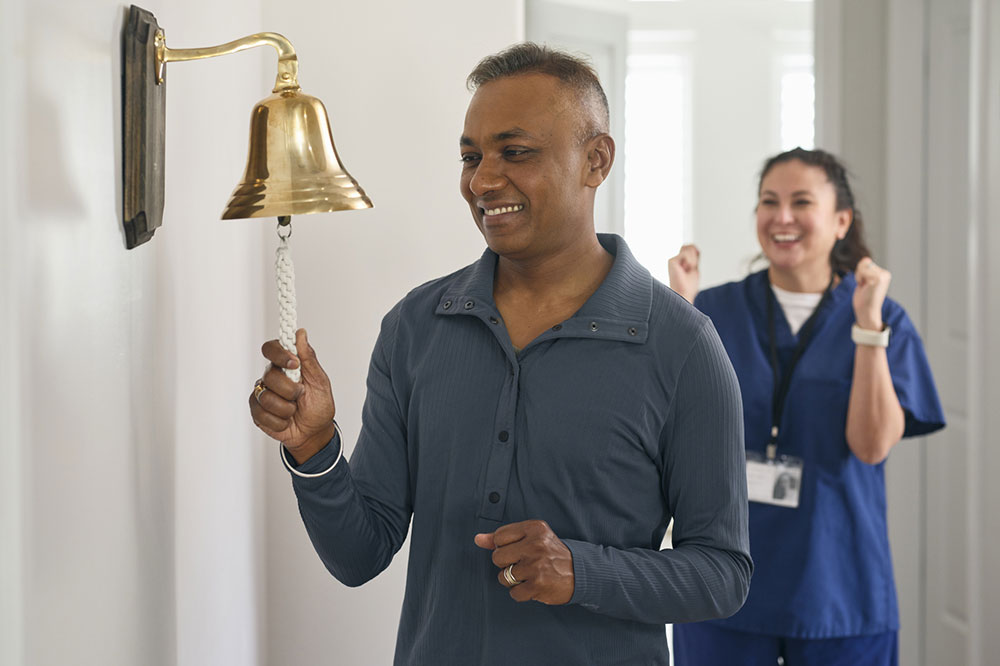 An man rings the bell in a treatment office to signify the end of his chemotherapy treatment office, as his patient navigator cheers in celebration.