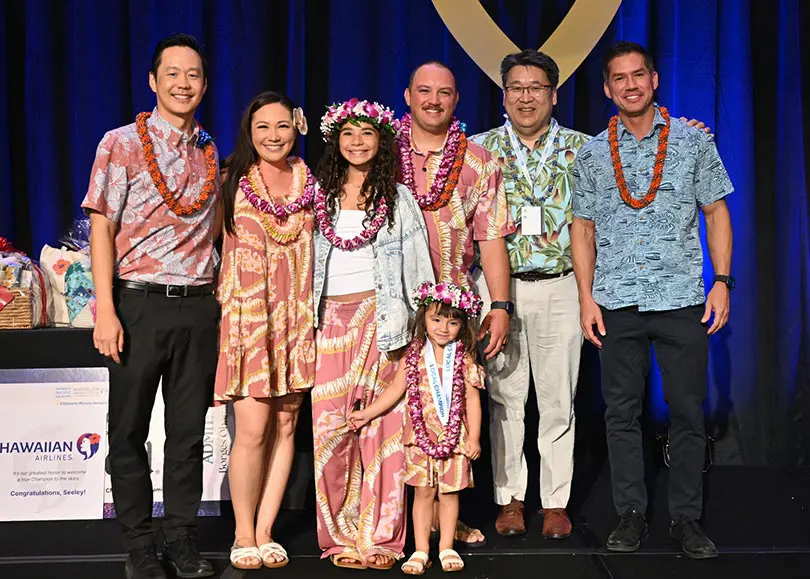 A group of people wearing floral attire and leis posing and smiling on stage in front of the CMN balloon logo.