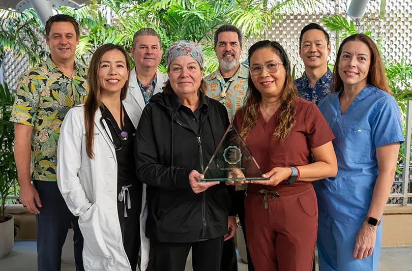 A group of eight healthcare professionals, smiling, pose with a triangular award. They wear diverse uniforms amid lush green plants, conveying a celebratory mood.