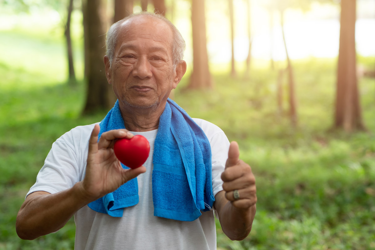 An elderly man with a sports towel draped over his shoulders standing outside in a wooded area holding a small felt heart. 
