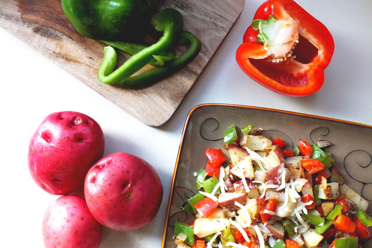 A potato hash sits on a plate near red potatoes and bell peppers.