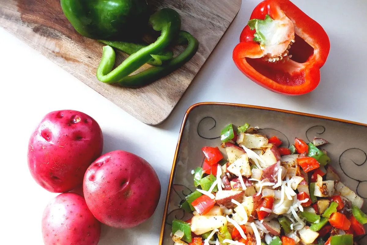 A potato hash sits on a plate near red potatoes and bell peppers.