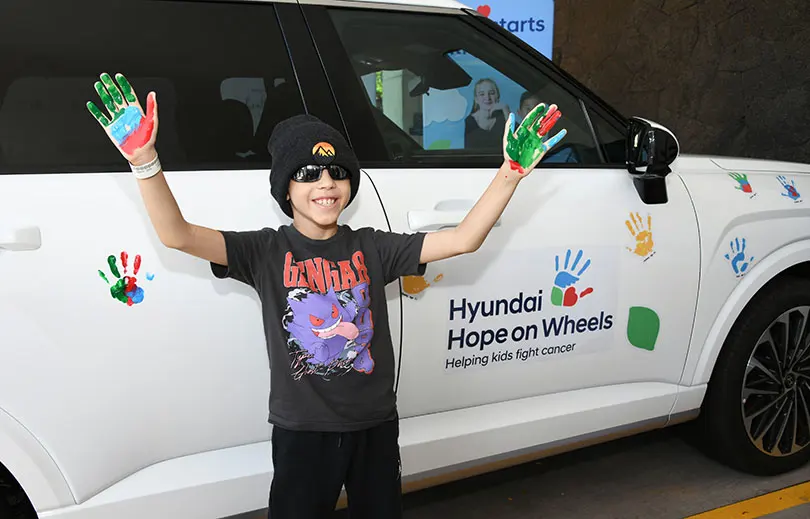 Young child with colorful paint on their hands standing in front of the Hyundai Hope on Wheels vehicle, decorated with colorful handprints.