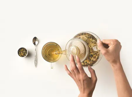 Hands pour a cup of tea with a spoon and container of loose tea nearby.
