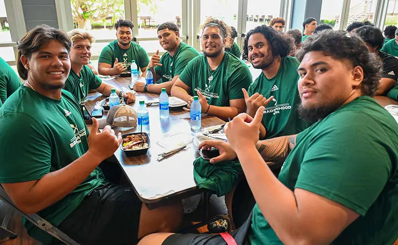 Players from the Rainbow Warrior football team gathered at a table, smiling, and enjoying lunch.