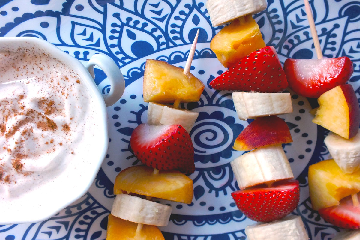 Peaches, bananas and strawberries alternate on skewers next a yogurt dip on a blue and white plate.