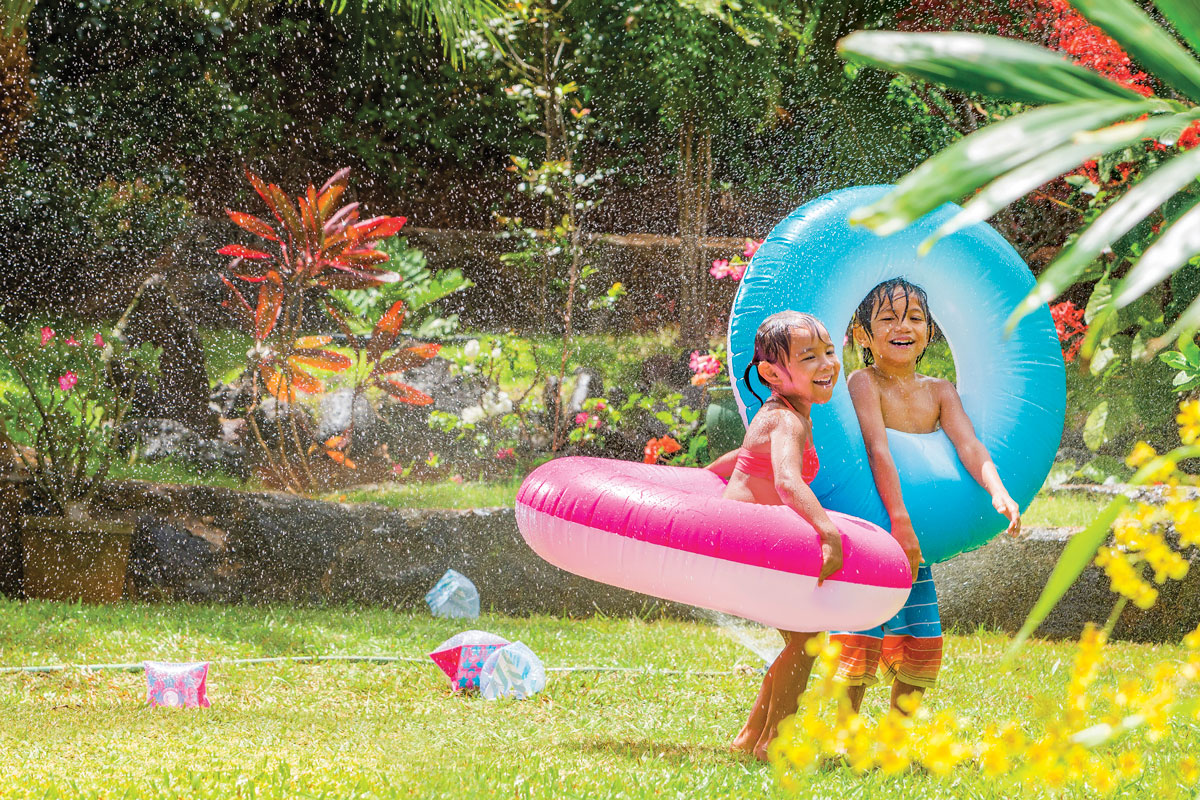 Two kids playing in the yard with a sprinkler and blow up pool toys.
