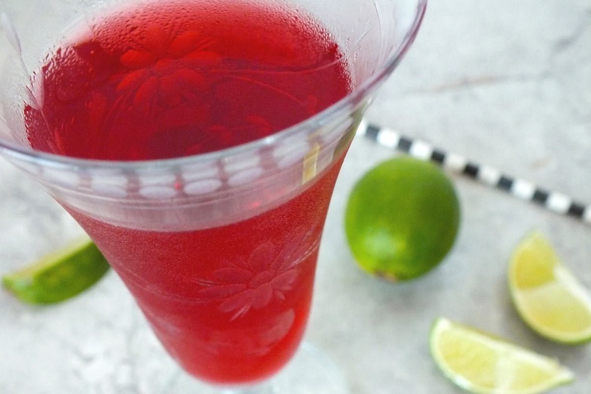 A bright red drink in a clear etched glass with limes and a black and white straw in the background.
