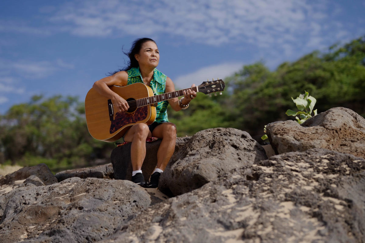 woman holds guitar while sitting on rocks, overlooking a beach.