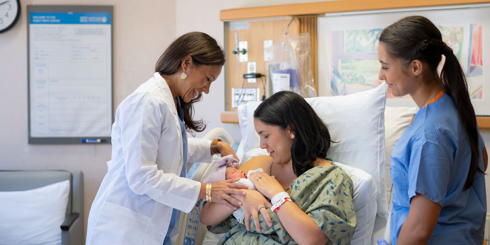 An OB-GYN stands over a mother and her newborn sitting in a bed in a delivery room at Kapiolani Medical Center and smiles, while a nurse stands on the opposite side of the bed and smiles.