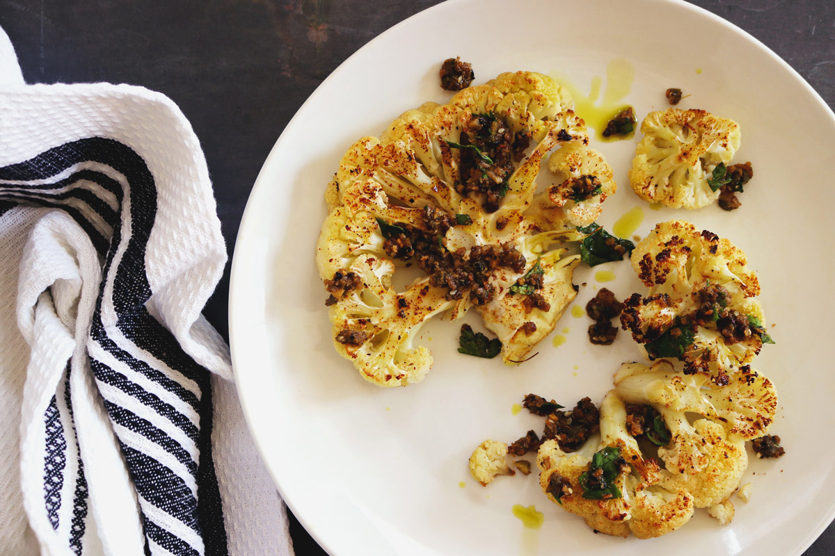 A plate of roasted Cauliflower Steaks drizzled with Spicy Chimichurri Sauce and a white-and-blue-striped napkin sit on a slate counter top.