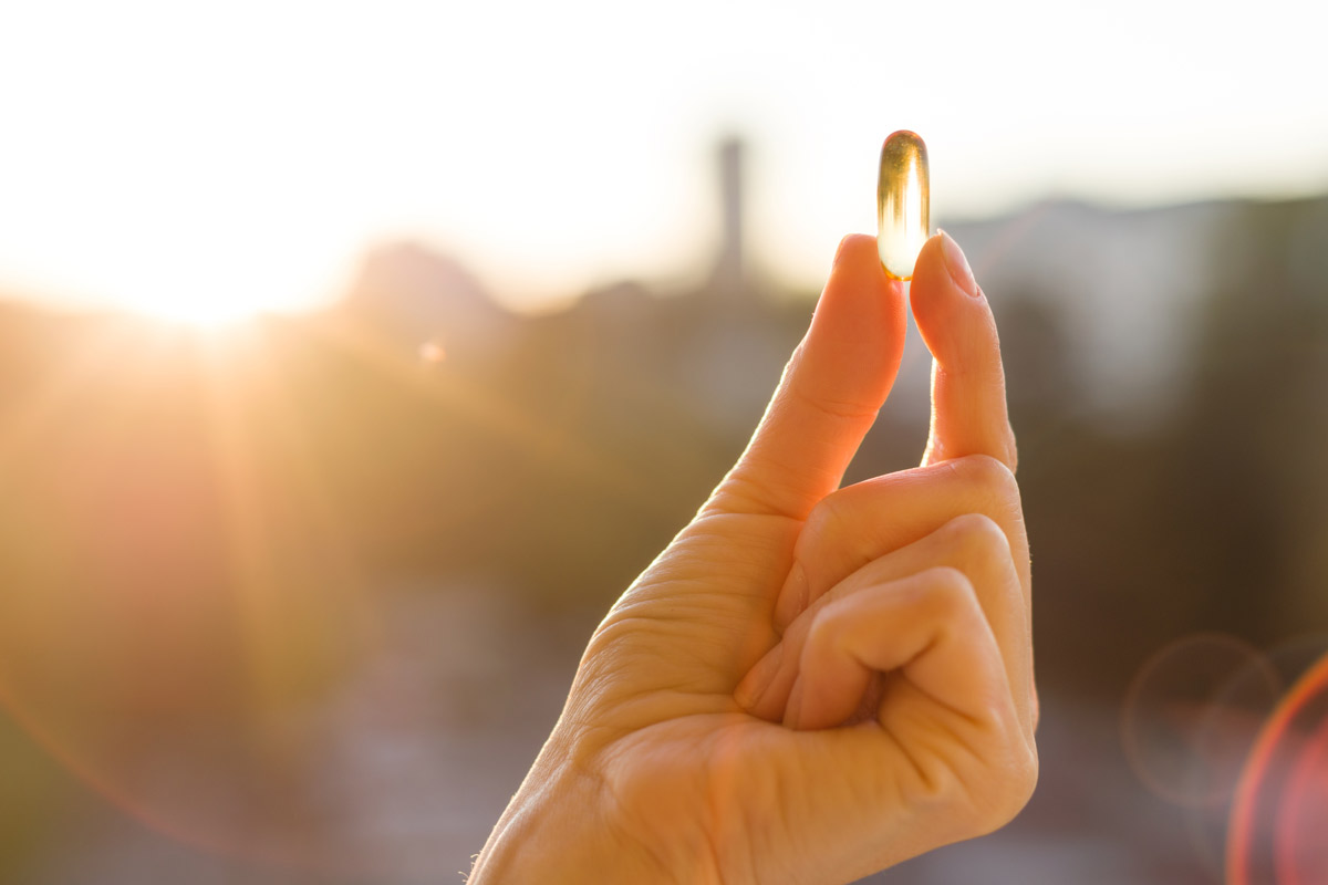 A hand holding up a vitamin D pill against a sunset.