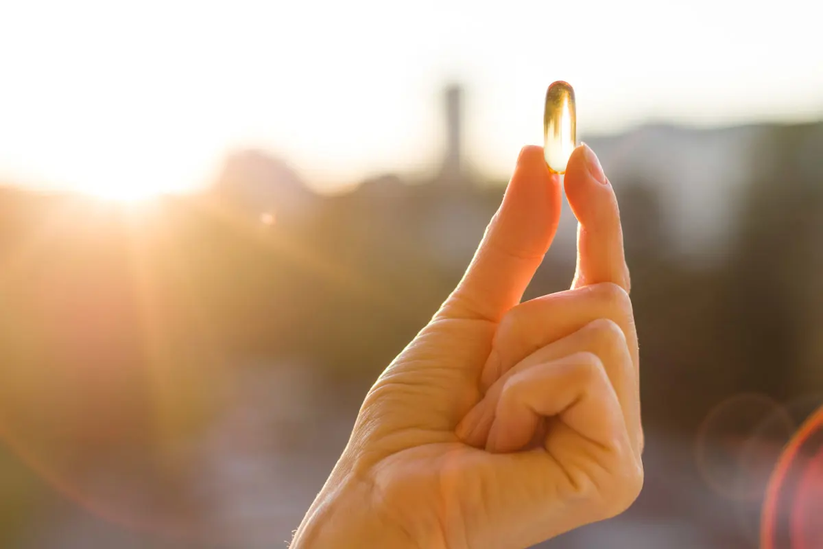 A hand holding up a vitamin D pill against a sunset.