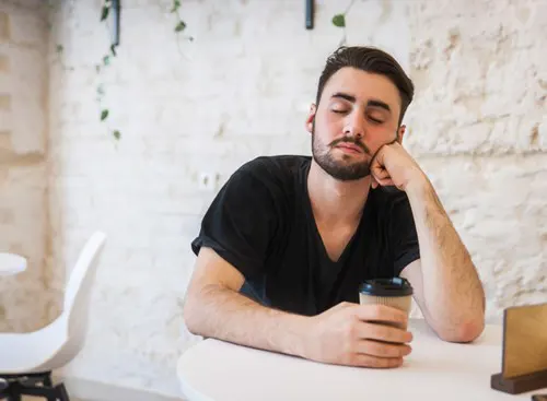 A man, with eyes closed, sits at a table with a cup of coffee.