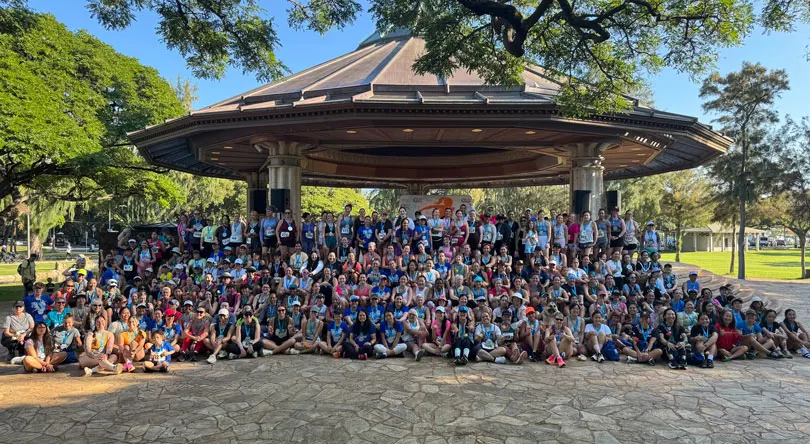 A large group of people gathered in front of a gazebo in a sunny park. Vibrant trees behind them. The atmosphere is cheerful and communal.